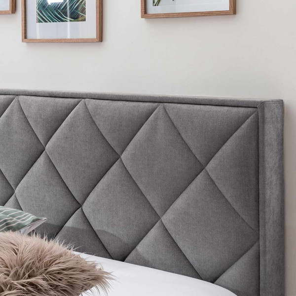 Boxspringbett mit Rautenmuster steht auf einem hellen Boden. Es hat graues Polster und weiße Bettwäsche. Eine Decke liegt auf einem Kissen, und im Hintergrund sind Pflanzenbilder und ein grüner Wandbelag sichtbar.
