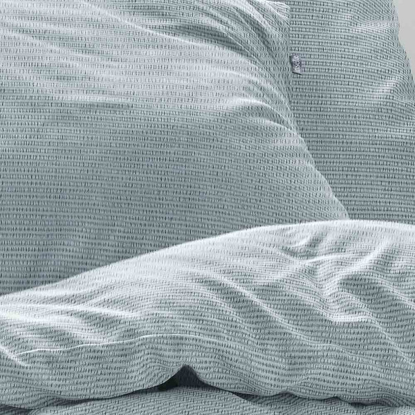 Zart gemusterte Irisette Seersucker-Bettwäsche in Hellblau liegt flach. Kissenbezug und Bettlaken sind ordentlich gefaltet und zeigen die textile Struktur und das dezente Design.