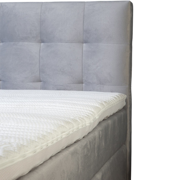 Boxspringbett mit grauem Stoffbezug steht auf einem Boden. Die Matratze hat einen Topper und ist in einer geschlossenen Position. Das Bett hat ein gepolstertes Kopfteil mit quadratischer Steppung.