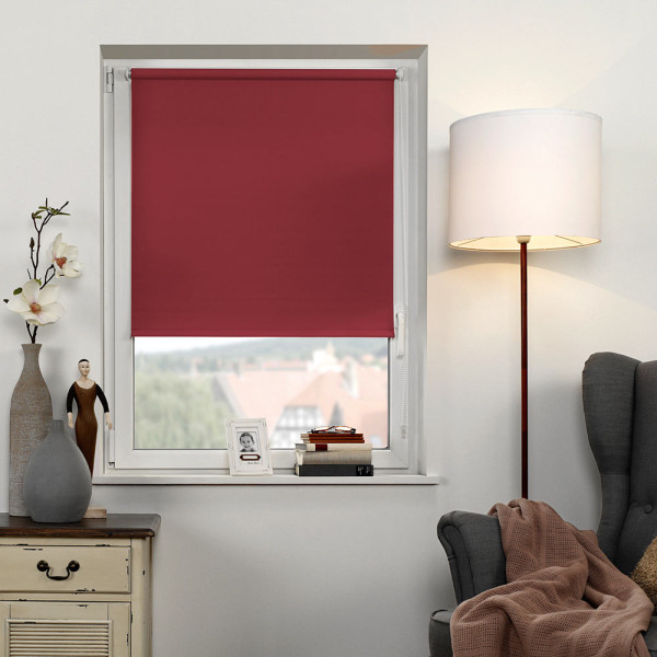 Rotes EasyFix Seitenzugrollo ALICIA hängt vor einem Fenster und bietet Lichtschutz. Eine Stehlampe leuchtet neben einem Sessel, während eine Vase mit Blumen und eine Figur auf einem Tisch stehen.