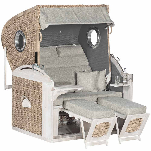 Strandkorb Fehmarn aus Pinien-Massivholz mit Rattangeflecht steht bereit. Er bietet zwei verstellbare Liegen und komfortable Kissen. Ein Getränk steht auf dem Tisch neben dem Korb.