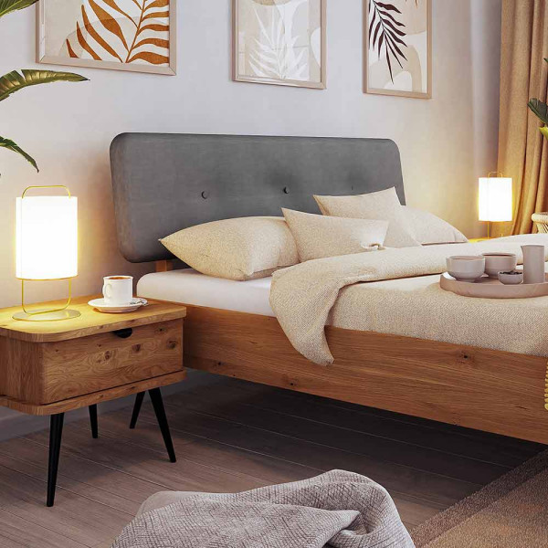 Modernes Bettgestell Kula aus Massivholz mit grauem Kopfteil, gelben Dekokissen und einer Decke in neutralen Tönen. Auf dem Bett befinden sich zwei Tassen und ein Tischgestell.