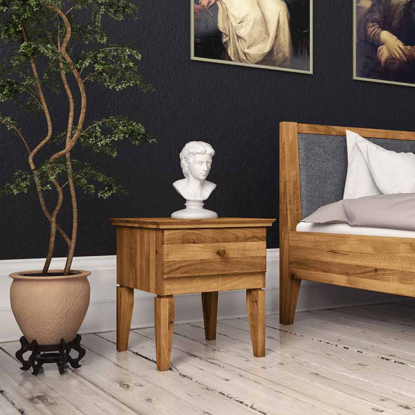 Rustikaler Nachttisch aus Holz steht neben einem Bett. Darauf befindet sich eine weiße Büste, während ein Topf mit einer Pflanze im Hintergrund steht. Die Wand ist dunkel gestrichen.