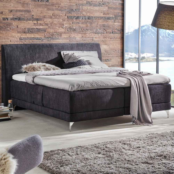 Elektrisch verstellbares Boxspringbett in Grau mit einem grauen Kopfteil und hellen Kissen. Eine Decke und ein Dekokissen liegen auf der Matratze. Ein Holzofenfenster zeigt eine LED-Lichtstimmung an.