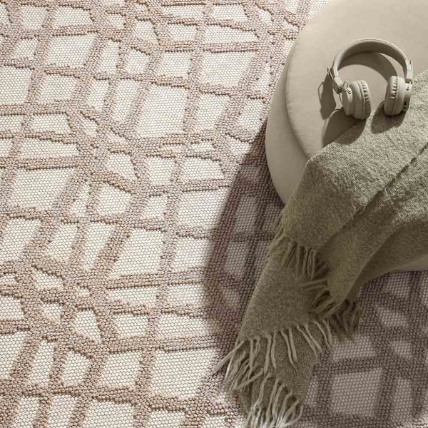 Hellbeiger Kurzflor-Teppich mit Hoch-Tief-Struktur liegt auf einem Holzfußboden. Ein runder Hocker mit einer Decke steht auf dem Teppich, während Tageslicht durch große Fenster strömt.