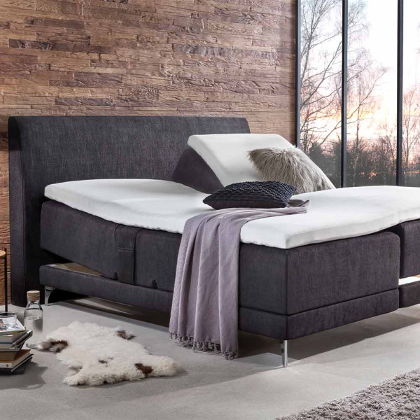 Elektrisch verstellbares Boxspringbett in Grau mit einem grauen Kopfteil und hellen Kissen. Eine Decke und ein Dekokissen liegen auf der Matratze. Ein Holzofenfenster zeigt eine LED-Lichtstimmung an.