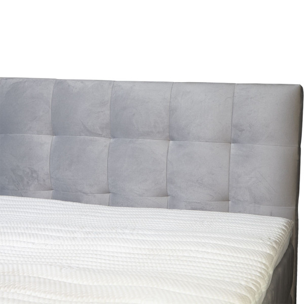 Boxspringbett mit grauem Stoffbezug steht auf einem Boden. Die Matratze hat einen Topper und ist in einer geschlossenen Position. Das Bett hat ein gepolstertes Kopfteil mit quadratischer Steppung.