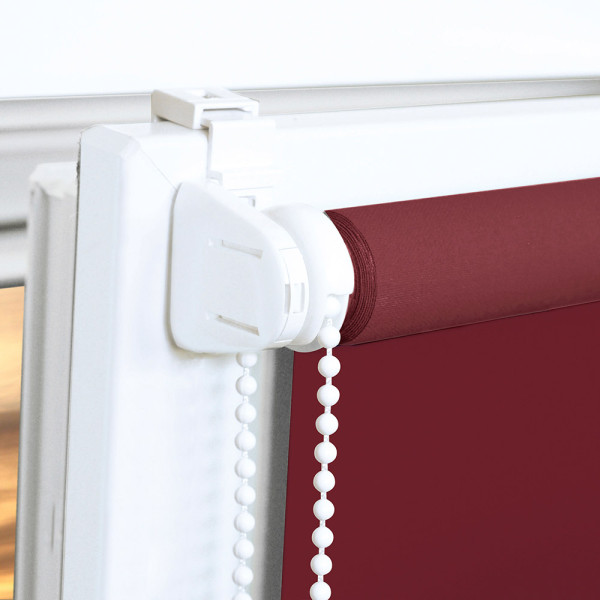 Roote in einem modernen Wohnraum ist ein rotes EasyFix Seitenzugrollo, das geschlossen ist. Eine Stehlampe beleuchtet einen gemütlichen Lesesessel neben einem Beistelltisch mit Blumen und einer Figur.