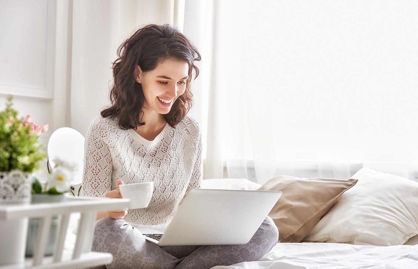 Junge Frau sitzt auf dem Bett und lächelt, während sie eine Tasse Kaffee hält und auf ihren Laptop schaut. Helle Vorhänge und ein Blumenarrangement sind im Hintergrund sichtbar.