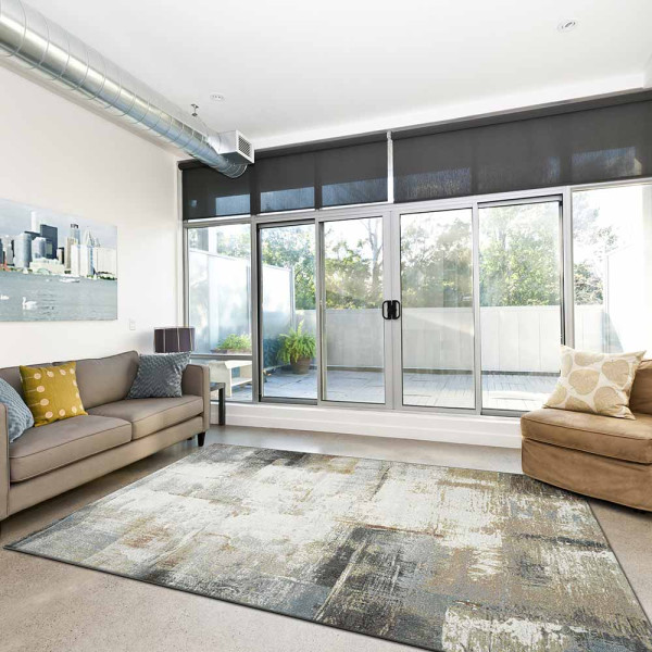 Grauer Kurzflor-Teppich Argentum 63723 mit abstraktem Muster in verschiedenen Grautönen und leicht goldfarbenen Akzenten liegt auf einem hellen Untergrund. Er vermittelt ein modernes Ambiente.