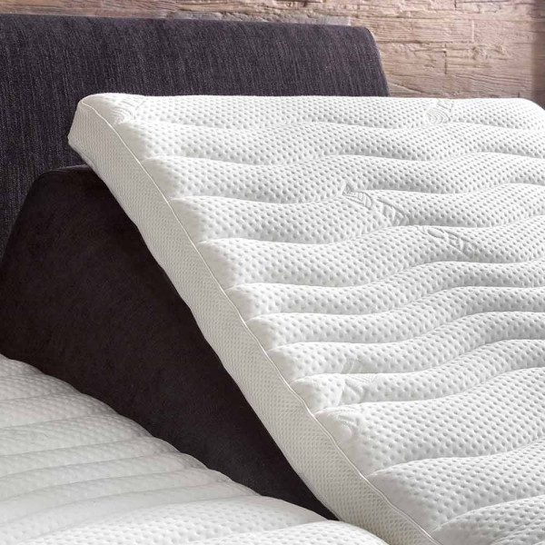 Elektrisch verstellbares Boxspringbett in Grau mit einem grauen Kopfteil und hellen Kissen. Eine Decke und ein Dekokissen liegen auf der Matratze. Ein Holzofenfenster zeigt eine LED-Lichtstimmung an.