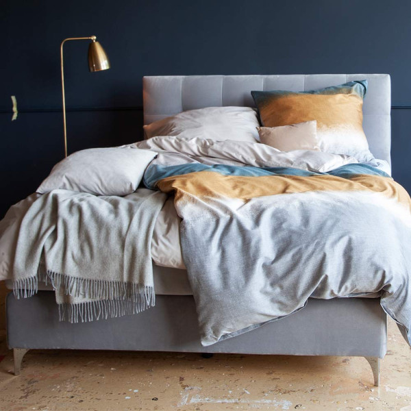 Boxspringbett mit grauem Stoffbezug steht auf einem Boden. Die Matratze hat einen Topper und ist in einer geschlossenen Position. Das Bett hat ein gepolstertes Kopfteil mit quadratischer Steppung.