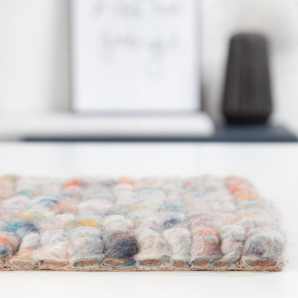 Bunte Teppichoberfläche aus Schurwolle mit einer sanften, strukturierten Optik. Die Farben reichen von Beige über Grau bis zu verschiedenen bunten Tupfern, was den Teppich vielseitig gestaltet.