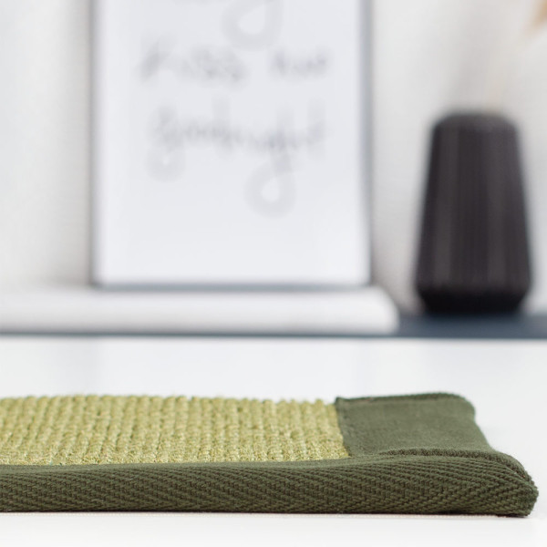 Sisal-Teppich in sanften Grüntönen hat eine strukturierte Oberfläche. An der Seite verläuft ein dunkles, gewebtes Band, das den Teppich einfängt. Ideal für individuelle Raumgestaltung.