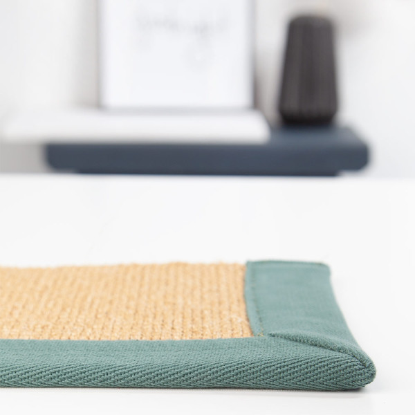 Beige Sisal-Teppichrand mit grünem Stoffbesatz ist sichtbar. Der Teppich zeigt eine naturbelassene Struktur und betont die nachhaltigen Materialien durch die harmonische Farbwahl.