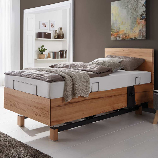 Komfortbett Lugano mit hochwertiger Matratze steht in einem modernen Raum. Es ist mit einer grauen Decke und Kissen dekoriert. Hinter dem Bett ist eine Wand mit Kunstwerk und einem Regal.