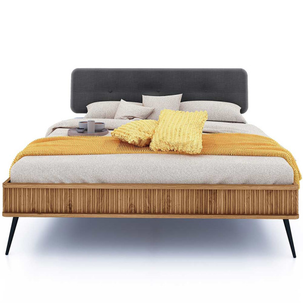 Modernes Bettgestell Kula aus Massivholz mit grauem Kopfteil, gelben Dekokissen und einer Decke in neutralen Tönen. Auf dem Bett befinden sich zwei Tassen und ein Tischgestell.
