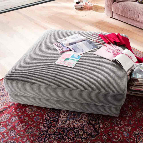 Grauer Hocker in Feincord-Optik mit verschiedenen Zeitschriften und einem roten Schal darauf. Er steht auf einem bunten Teppich in einem hellen Raum.