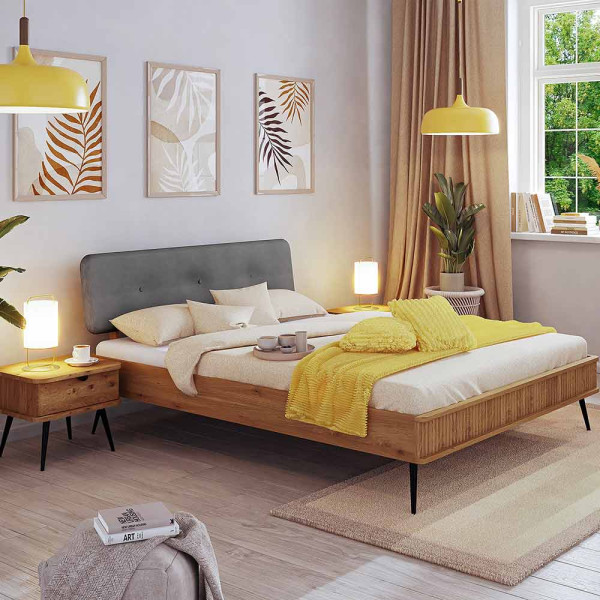 Modernes Bettgestell Kula aus Massivholz mit grauem Kopfteil, gelben Dekokissen und einer Decke in neutralen Tönen. Auf dem Bett befinden sich zwei Tassen und ein Tischgestell.