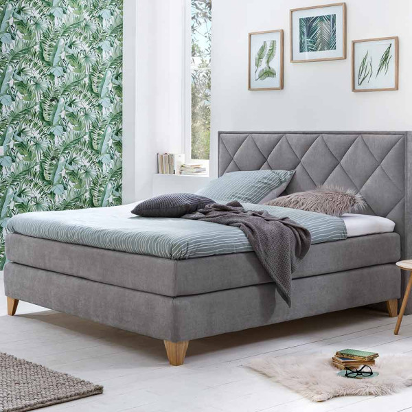 Boxspringbett mit Rautenmuster steht auf einem hellen Boden. Es hat graues Polster und weiße Bettwäsche. Eine Decke liegt auf einem Kissen, und im Hintergrund sind Pflanzenbilder und ein grüner Wandbelag sichtbar.