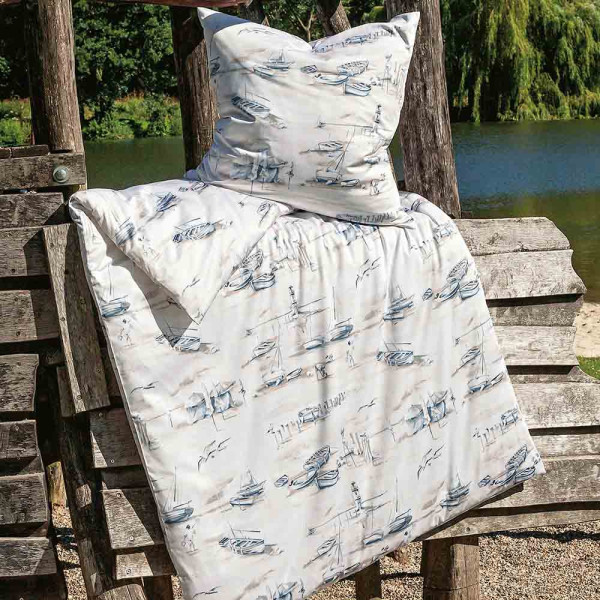 Bettwäsche mit maritimem Design zeigt Boote, Leuchttürme und Möwen in sanften Blau- und Grautönen. Die Garnitur umfasst einen Bettbezug und zwei Kissenbezüge, die sanft drapiert sind.