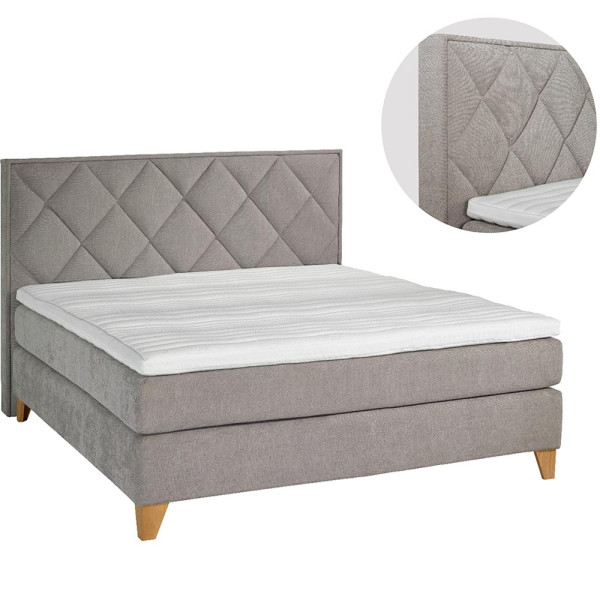 Boxspringbett mit Rautenmuster steht auf einem hellen Boden. Es hat graues Polster und weiße Bettwäsche. Eine Decke liegt auf einem Kissen, und im Hintergrund sind Pflanzenbilder und ein grüner Wandbelag sichtbar.